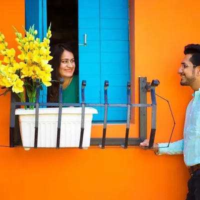 For this pre-wedding shoot, we used a colorful, vibrant setting to create a playful 'Romeo and Juliet' scene. The bright orange wall and blue window provide a high-contrast background that makes the couple's interaction pop.