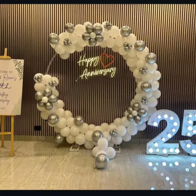 An elegant setup for a 25th wedding anniversary. The large circular balloon arch in white and silver, a custom neon sign, and big light-up "25" numbers create a sophisticated photo backdrop.