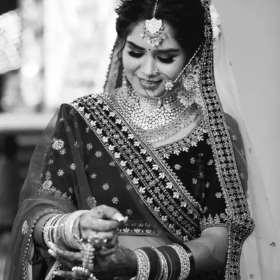 A timeless black and white bridal portrait. This image focuses on the bride's expression as she looks down at her kalire. Converting the photo to monochrome removes distractions, emphasizing the rich textures of her velvet lehenga and the intricate details of her jewelry.