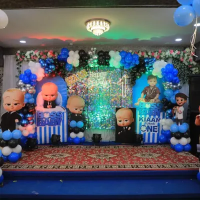 A full stage setup for a Boss Baby first birthday party, featuring multiple character cutouts, a silver shimmer backdrop, and themed balloon pillars in a large hall.