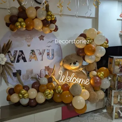 A chic and modern welcome home decor for baby Vaayu. The setup features a custom backdrop, a large plush teddy bear, and a sophisticated balloon arrangement in earthy tones with pops of metallic gold.