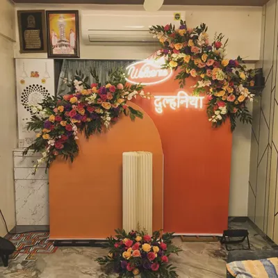 A "Welcome Dulhania" decoration for a new bride's arrival. This home setup features a warm orange backdrop, a custom neon sign, and beautiful floral arrangements.