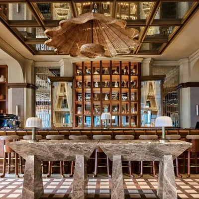 The iconic pour bar, with its monolithic marble tables and a dramatic, shell-like rattan chandelier. The mirrored ceiling reflects the scene, amplifying the energy and maximalist aesthetic of the space.