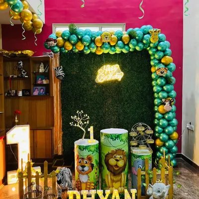 A full view of a jungle safari first birthday party setup in a home. The decoration features a lush green grass wall, a gold and green balloon arch with animal head foils, themed pedestals, and a large light-up number '1'.