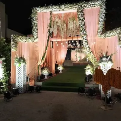 A luxurious nighttime wedding entrance featuring soft peach drapes, a full arch of white flowers, and elegant chandeliers. Spotlights illuminate the path, creating a grand and welcoming feel.