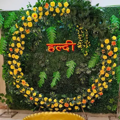 A modern take on Haldi decor, this setup features a circular frame covered in yellow flowers against a dense green foliage wall. The floral 'Haldi' lettering in the center adds a personalized touch to this fresh and stylish photo backdrop.