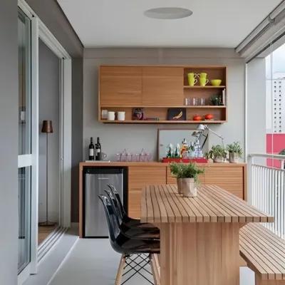 A minimalist and highly functional balcony bar setup. This design features clean wooden cabinetry with open shelving, a built-in mini-fridge, and a matching dining table with a bench. It's a practical solution for creating an al fresco dining experience.
