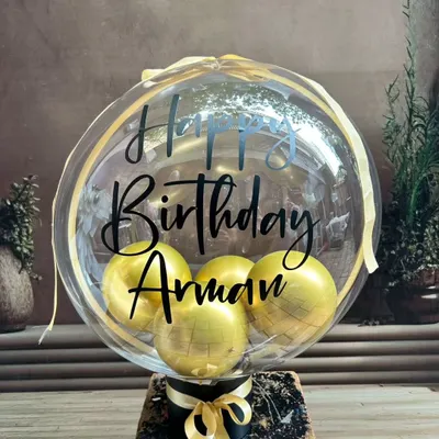 A personalized balloon bouquet featuring a large transparent balloon with a custom 'Happy Birthday' message and smaller gold balloons inside. It's a perfect gift or a centerpiece for a celebration.