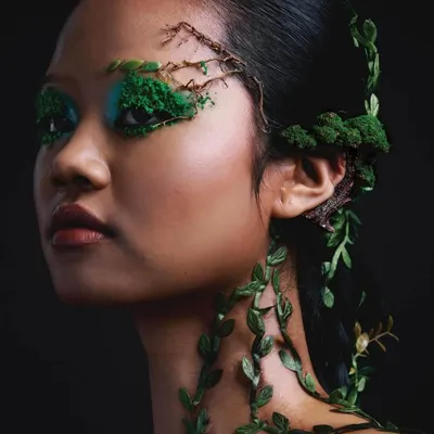 A powerful three-quarter shot against a stark black background, highlighting the intricate natural details in the makeup and hair. The model wears vines that creep up her neck and a miniature tree ear cuff, blending fashion with the earth.