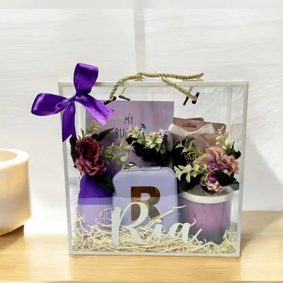 This personalized gift for 'Ria' is presented in a stunning transparent acrylic bag with a purple ribbon. It features a custom initial on a travel case and other curated items in a matching lavender theme.