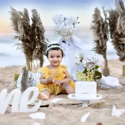 A beautiful first birthday setup on the beach, with a baby girl in a yellow dress sitting beside her cake, with the ocean in the background.