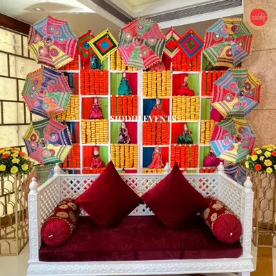 We created this joyful Haldi backdrop using colorful Gujarati umbrellas, marigold garlands, and traditional Rajasthani dolls, all set against a white diwan with rich red cushions.