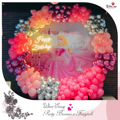A fairytale party for Princess Katherine! This enchanting setup features a custom princess backdrop framed by a lush organic balloon garland in shades of pink, purple, and silver, all lit by a 'Happy Birthday' neon sign.