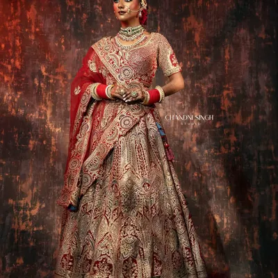 A full-length bridal portrait showcasing a magnificent red and gold lehenga. The lighting is sculpted to highlight the rich embroidery and create a regal, statuesque presence.