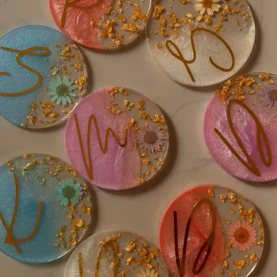 A collection of my custom resin initial coasters. Each piece is unique, with different color combinations like pink, blue, and white, and all are adorned with real dried flowers and sparkling gold leaf.