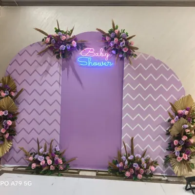 A modern boho baby shower backdrop in lavender and white. This design features chevron-patterned panels, a 'Baby Shower' neon sign, and beautiful arrangements of purple flowers and dried golden leaves.