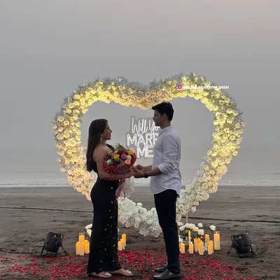 A couple enjoys their special moment in front of a heart-shaped floral arch on the beach. This complete proposal package includes the illuminated arch, neon sign, and romantic ground lighting to capture the perfect "yes".