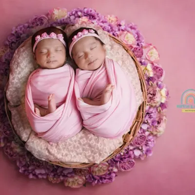 The perfect pair. Two newborn twin girls swaddled in pink, sleeping side-by-side in a basket surrounded by purple flowers. This is a classic and heartwarming twin portrait.
