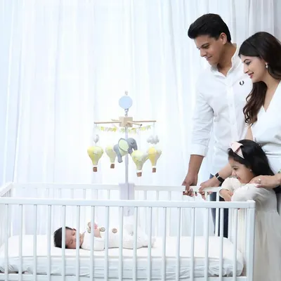 A heartwarming lifestyle scene where parents and an older sister gather around the crib to watch over the family's newest member. The all-white outfits create a clean, serene atmosphere.