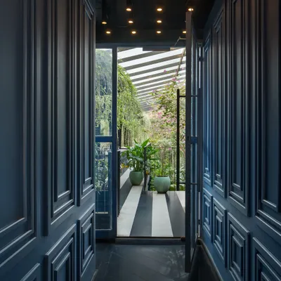 A doorway framed by deep blue paneled walls offers a glimpse from a moody corridor into the bright, plant-filled garden court. This transition highlights the deliberate contrast I create between different zones to evoke distinct emotional responses.