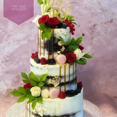 A three-tiered semi-naked cake for a proposal, topped with a "She Said Yes" acrylic sign. This rustic yet elegant design is adorned with fresh red and white roses, berries, and a caramel drip.