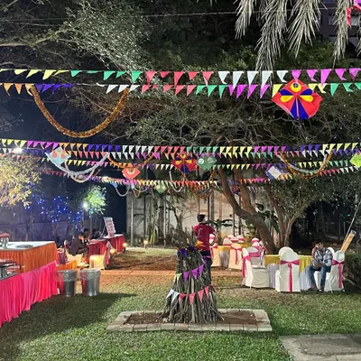An outdoor Lohri celebration at night. The garden is decorated with colorful flags, kites, and traditional hangings, with a bonfire set up in the center.