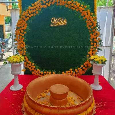 A beautiful Haldi ceremony setup featuring a circular marigold wreath against a green grass wall backdrop. A custom neon "Haldi" sign adds a modern touch, while a traditional golden lotus-shaped basin for the rituals sits in the foreground on a red carpet.