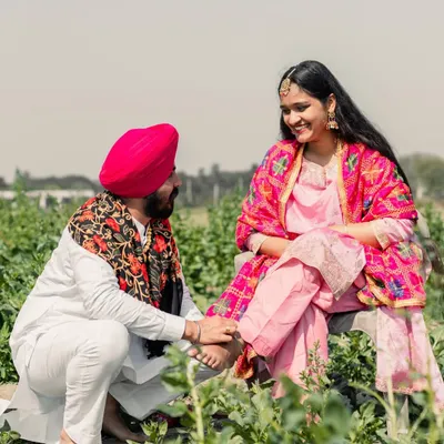 A beautiful, clear shot of a groom-to-be tenderly placing an anklet on his partner's foot in a lush green field, a classic Punjabi romantic gesture.