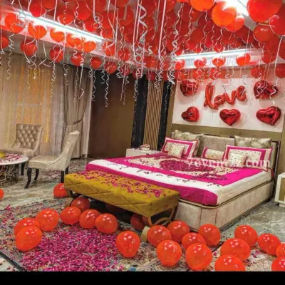 A full room transformation for a first night celebration. This setup features hundreds of red balloons with ribbons covering the ceiling, a dense scattering of balloons on the floor, and a bed adorned with rose petals for a classic romantic feel. The 'LOVE' and heart-shaped foil balloons on the wall complete the look.