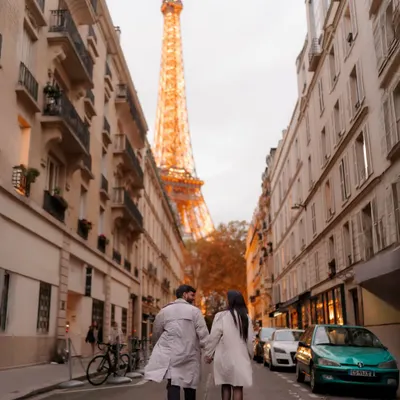 A classic Paris street scene with a romantic twist. The couple walks hand-in-hand away from the camera down a quintessential Parisian street, with the Eiffel Tower perfectly framed in the distance at golden hour.