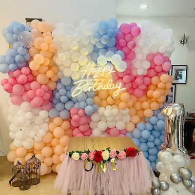 A magnificent floor-to-ceiling balloon wall in a mix of pastel colors, with a central neon 'Happy Birthday' sign. The setup is completed with a tulle-draped table for the cake.