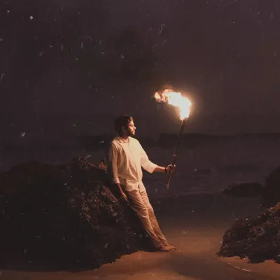 A striking solo portrait of the groom-to-be, leaning against a rock on the beach at night while holding a flaming torch. The firelight creates dramatic shadows, resulting in a powerful and moody pre-wedding shot.