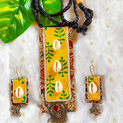A vibrant yellow fabric panel, hand-painted with green leaf motifs and adorned with cowrie shells, is the centerpiece of this necklace. It's layered over a traditional Kalamkari print and finished with an oxidized silver charm. This is a perfect example of my layered, mixed-media style.