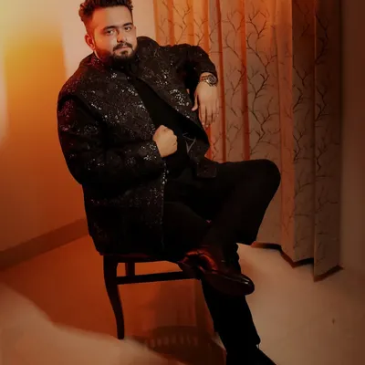 A confident, full-length portrait of the groom seated, showcasing his stylish black sherwani against a warm, lit background.