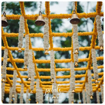 A close-up of a mandap ceiling, showcasing a beautiful grid of yellow and white chrysanthemum garlands with hanging temple bells. This detail adds sound, color, and tradition to the ceremony.