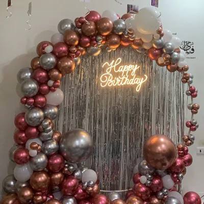 Our 'Happy Birthday' neon sign is the perfect centerpiece for this home setup, glowing against a silver fringe curtain and a metallic balloon arch.