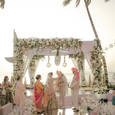 A stunning wide shot of a beach wedding ceremony at sunset. The couple stands under a magnificent floral mandap as the golden light kisses the horizon.