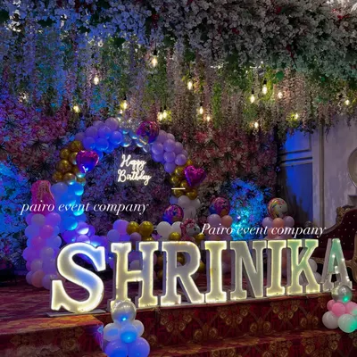 A beautiful stage setup featuring large, illuminated marquee letters spelling out "SHRINIKA." The letters are the main attraction, set against a backdrop of hanging flowers and lights.