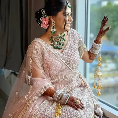 A picture of pure bridal grace. This look features soft, romantic makeup with a pop of pink in the floral bun, perfectly complementing her blush lehenga and emerald jewellery for a timeless and elegant feel.