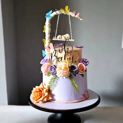 A magical baby shower cake in a peach and lavender color scheme. The centerpiece is a hand-sculpted, edible baby sleeping in a swinging bassinet, surrounded by flowers and butterflies.