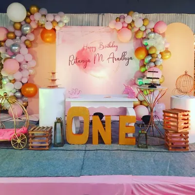 A beautiful first birthday setup for Ritanya, featuring a soft, watercolor-style backdrop. The stage is decorated with a pastel balloon garland, gold shimmer panels, and large marquee letters spelling 'ONE', creating an elegant and celebratory focal point.