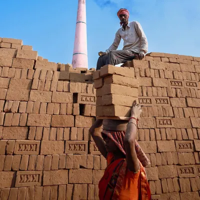A woman carries a stack of bricks on her head while a man watches from atop a brick wall, a colorful scene depicting the daily labor at a kiln in Bihar.