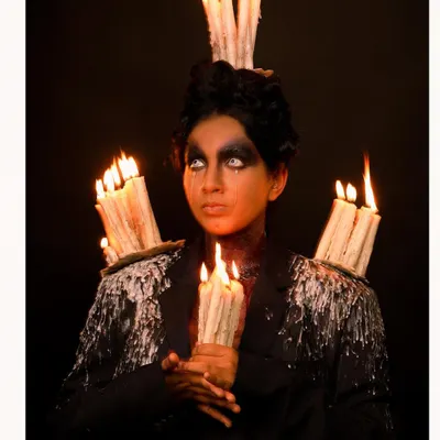 A dark fantasy portrait featuring a model with dramatic makeup, holding lit candles, and wearing a custom jacket adorned with dripping wax and more candles. This is an example of my highly conceptual and artistic work.