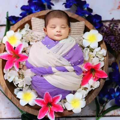 A sleeping newborn is wrapped in purple and white cloth, nestled in a wooden bowl adorned with beautiful, colorful flowers.