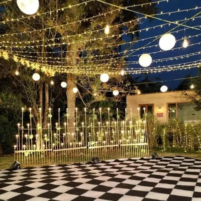 An outdoor Sangeet setup designed for a night of celebration. The checkerboard dance floor is surrounded by a fence of lights and set beneath a canopy of string lights and paper lanterns, creating a magical atmosphere.