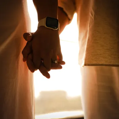 A close-up of the couple holding hands against the warm glow of the sun. The focus is on their intertwined fingers and the new engagement ring, symbolizing their connection.