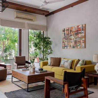 This living room seamlessly blends Tropical Modernism with Boho-Chic elegance. Large glass doors invite nature inside, while a lime-toned sofa and rich wooden finishes create a space filled with both calm and character.