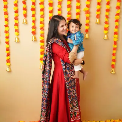 A mother in a beautiful red traditional outfit holds her son during a Ganesh Chaturthi celebration. The background is decorated with marigold garlands, creating a vibrant and festive scene.