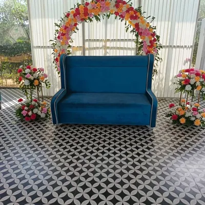 This engagement photo corner features a vibrant semi-arch of artificial flowers in shades of pink, orange, and yellow. The setup is completed with a stylish blue wingback sofa for the couple, making it a perfect spot for pictures.