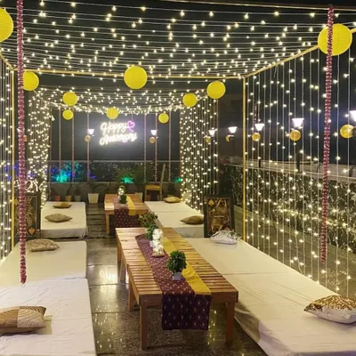 A stunning terrace anniversary setup featuring a canopy of fairy lights and yellow paper lanterns. The low wooden tables with floor seating create a cozy and intimate atmosphere for a group celebration.
